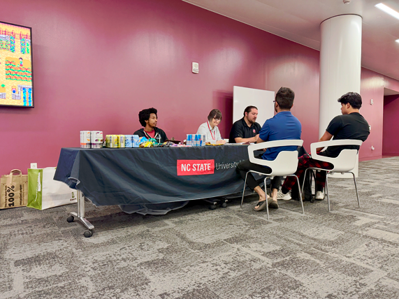 Three Libraries staff members sit at a table with energy drinks and snacks, talking to two students who are seated in front of the table.