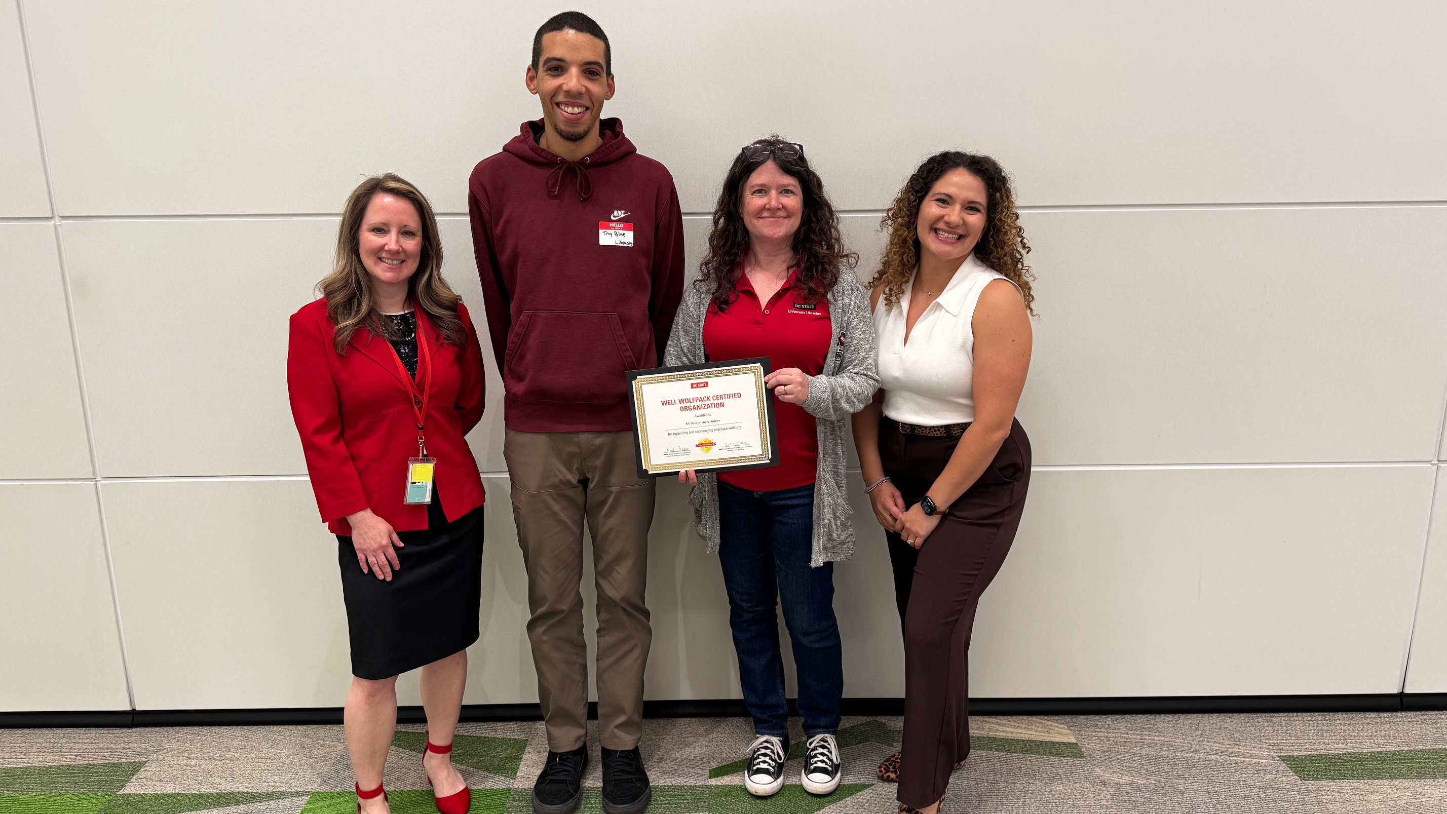 Four people stand against a wall for a group photo. One person holds a framed certificate.