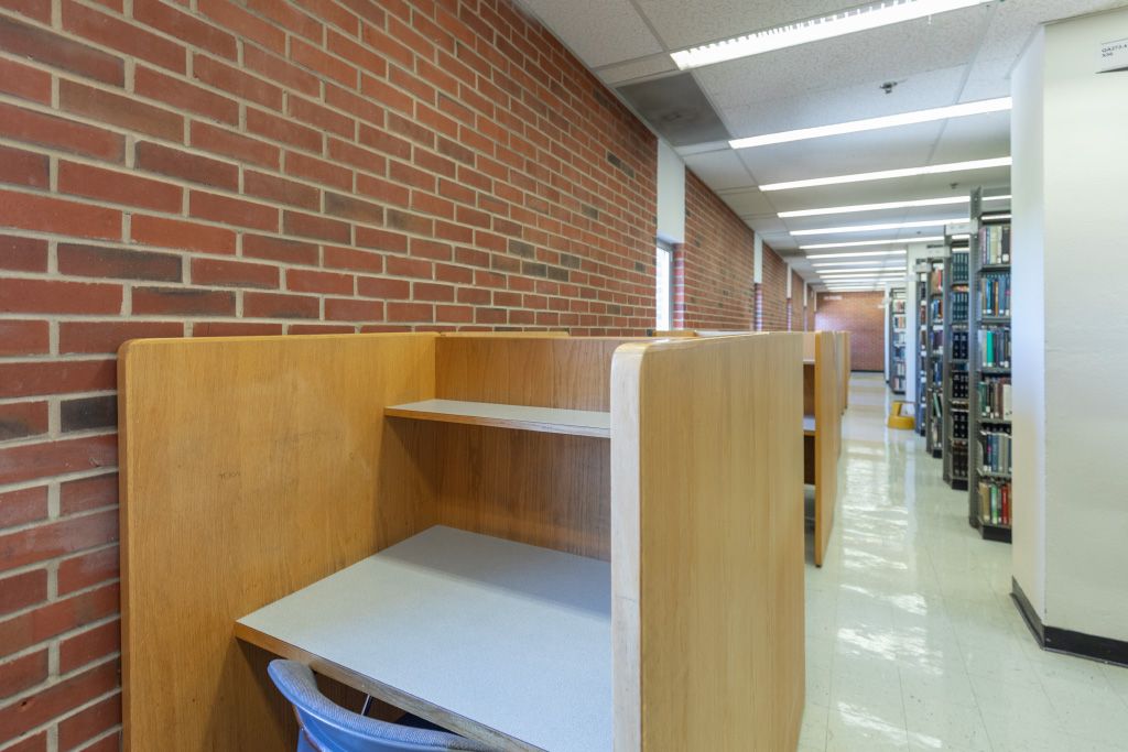 Individual carrels along a brick wall with exterior windows across the aisle from bookshelves.