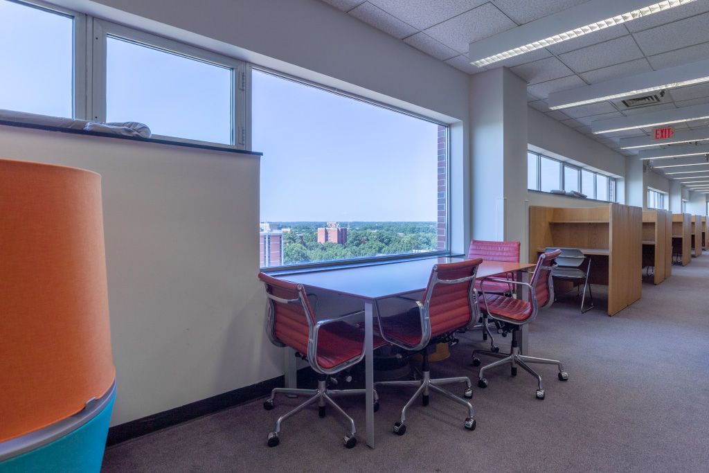 Large table with three task chairs overlooking campus through exterior window with individual carrels in the background.