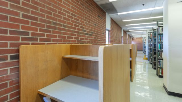 Individual carrels along a brick wall with exterior windows across the aisle from bookshelves.